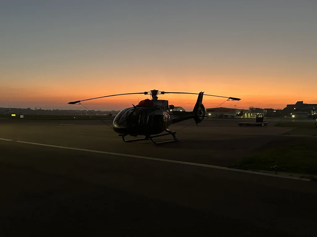 An Airbus helicopter parked on the apron in the dusk. Main rotor blades tied down.